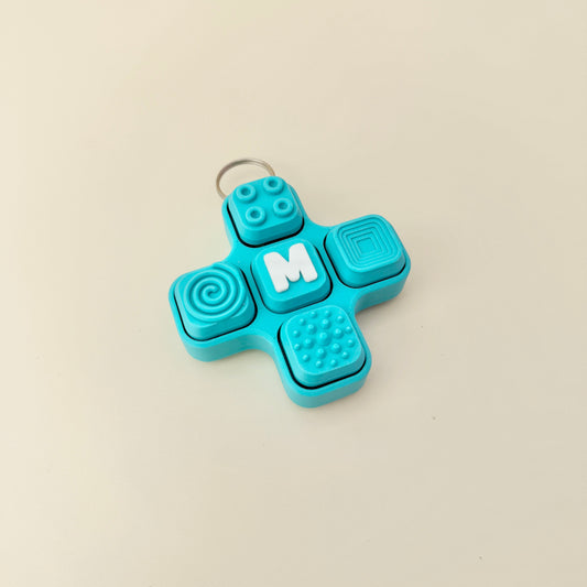 Blue fidget spinner keychain with textured surface and letter 'M' on a beige background