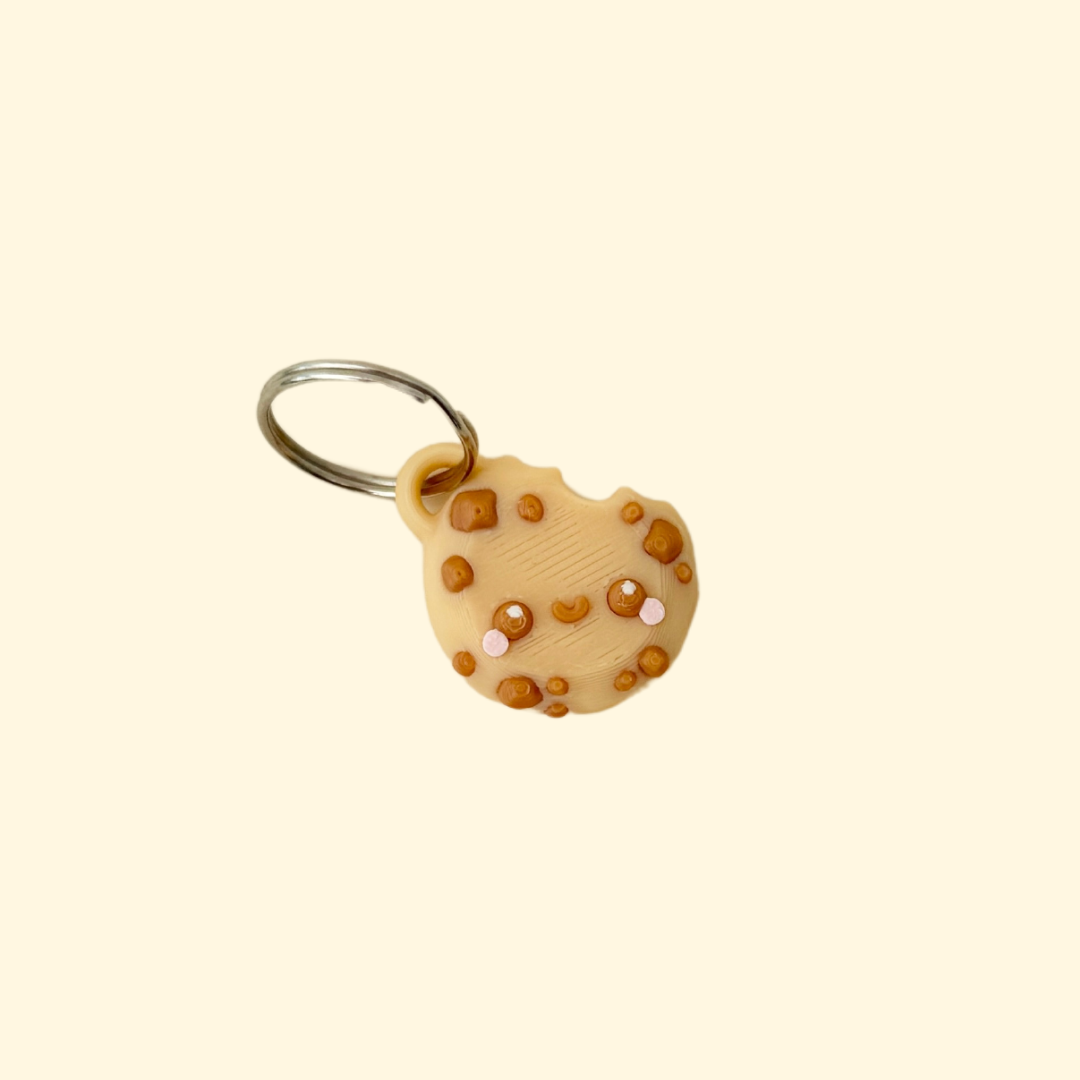 A small, beige chocolate chip cookie keyring with a bite taken out, featuring cute eyes, a smile, and pink cheeks.