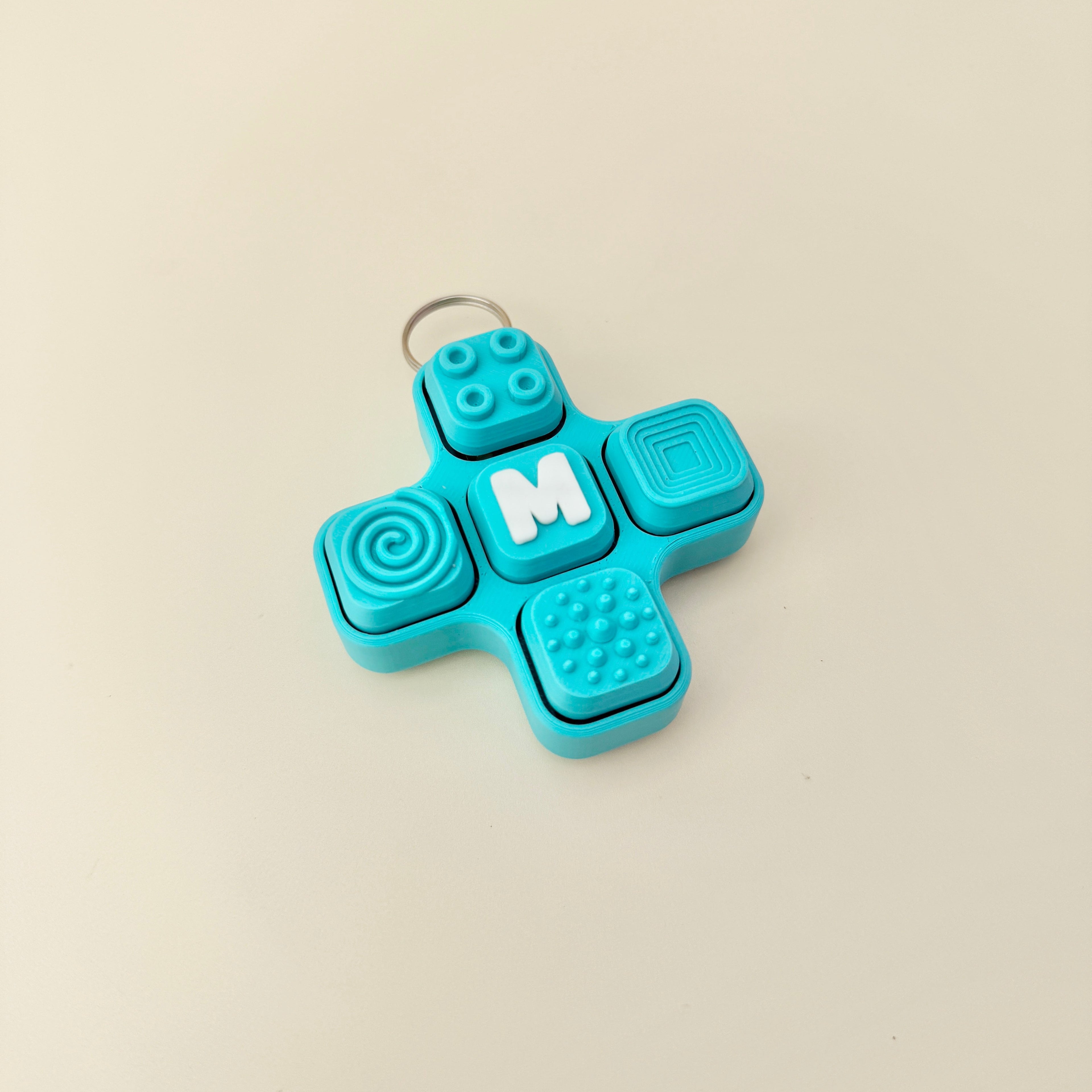 Blue fidget spinner keychain with textured surface and letter 'M' on a beige background