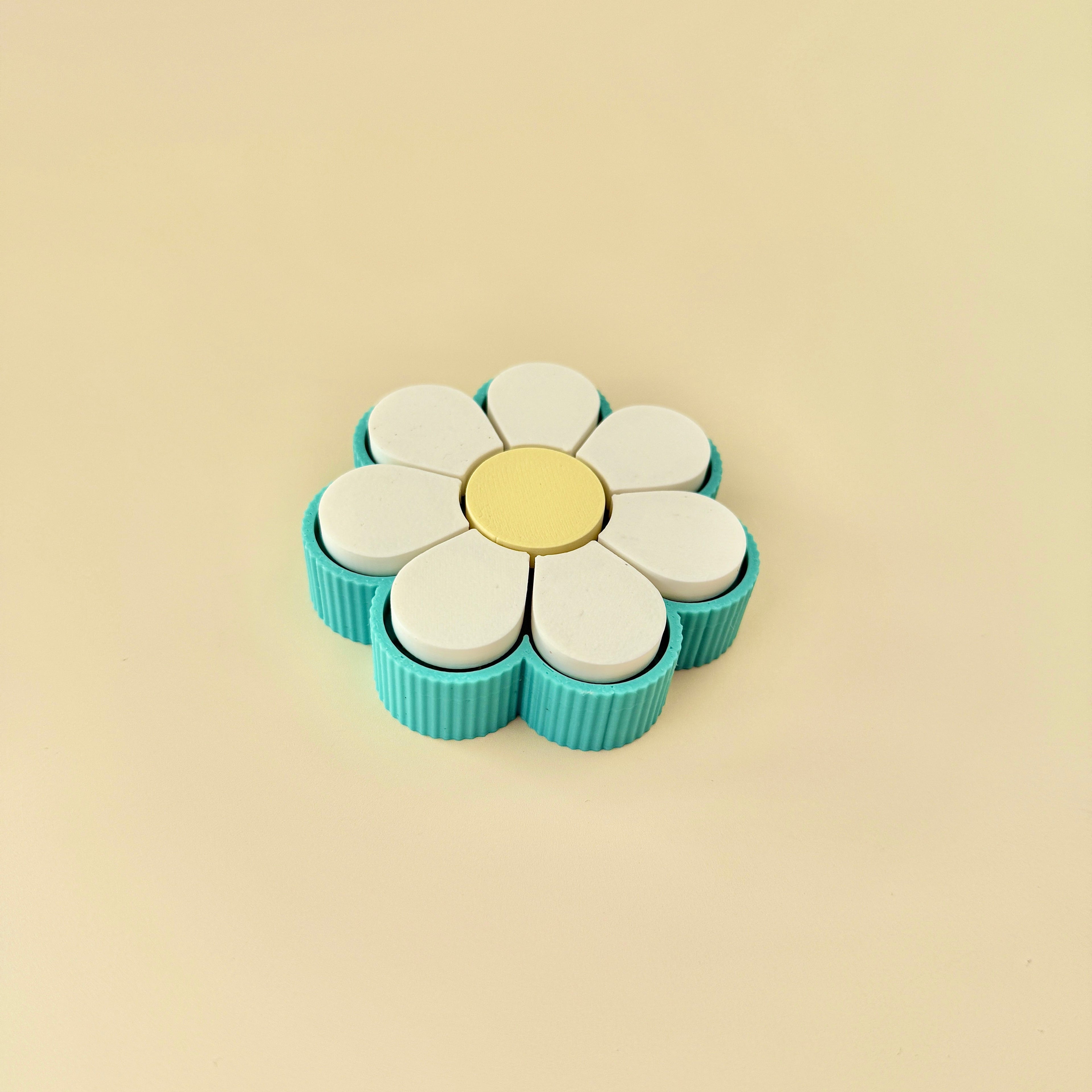 Daisy-shaped clicker featuring white petals, a yellow centre, and teal ridged outer grip, ideal for desk-based fidgeting.