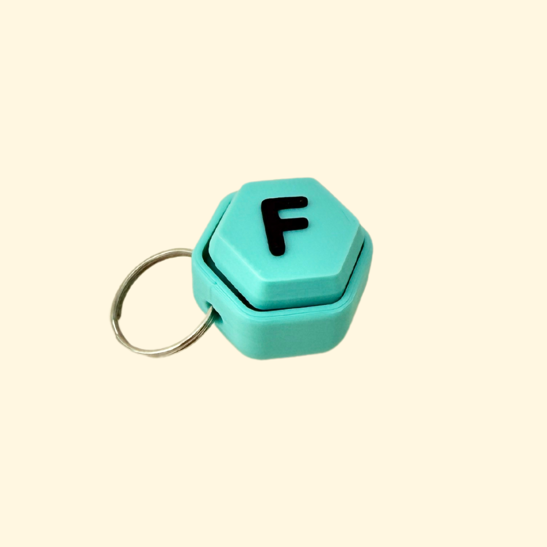 A teal hexagon-shaped fidget keyring featuring a raised black letter for personalised sensory play.