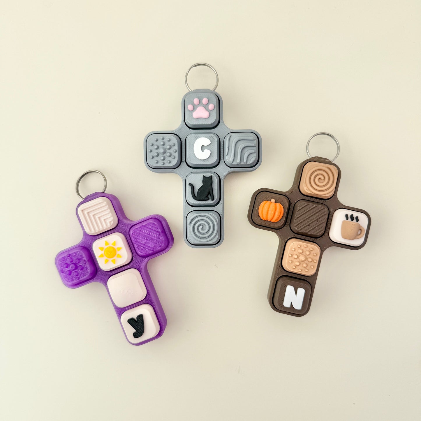 Three colorful cross-shaped fidget keychains with various designs on a beige background