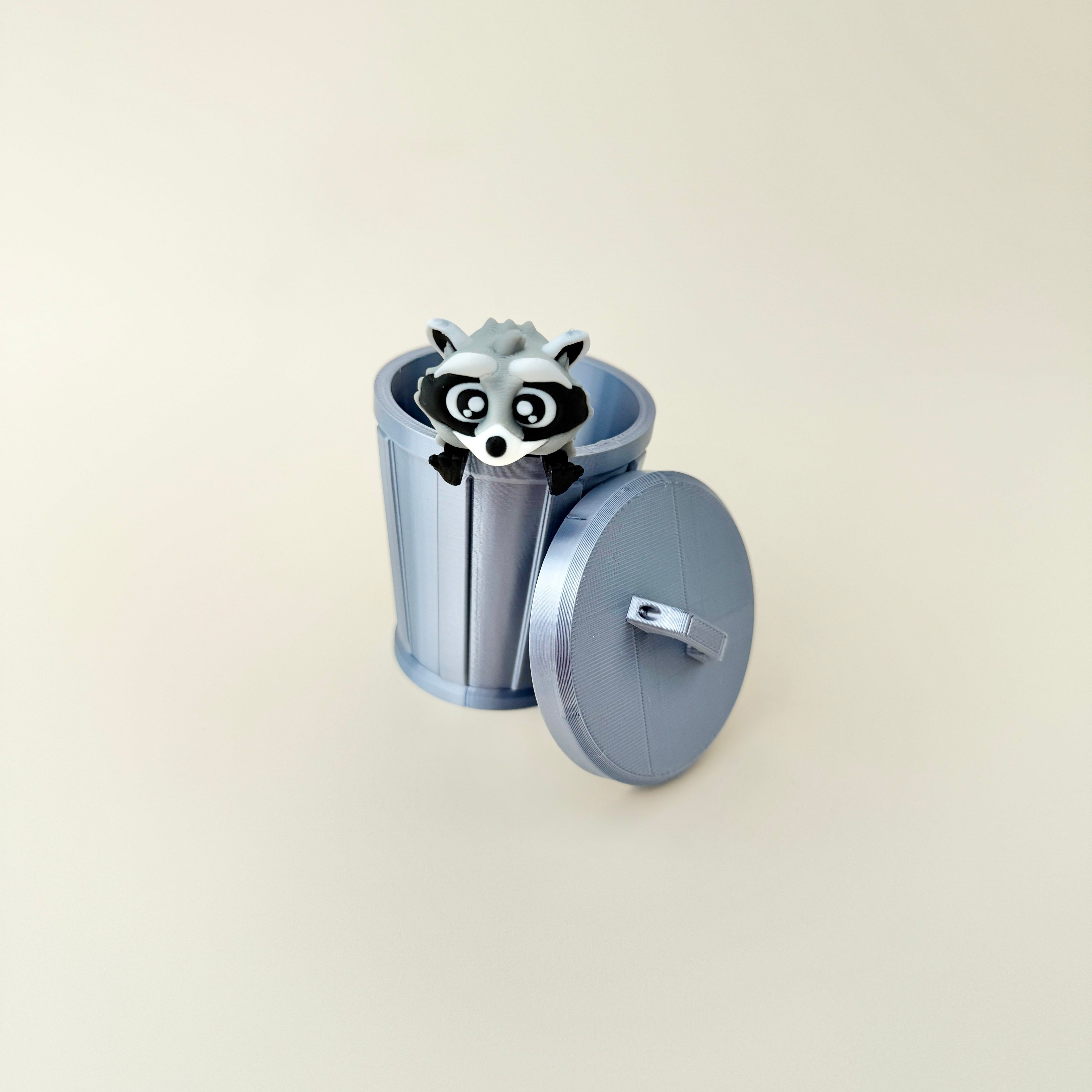 A smooth grey trash can with a removable lid, designed to fit the raccoon fidget inside.