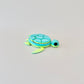 Green and blue turtle-shaped fidget clicker with moveable head, legs, and tail, designed for stress relief.