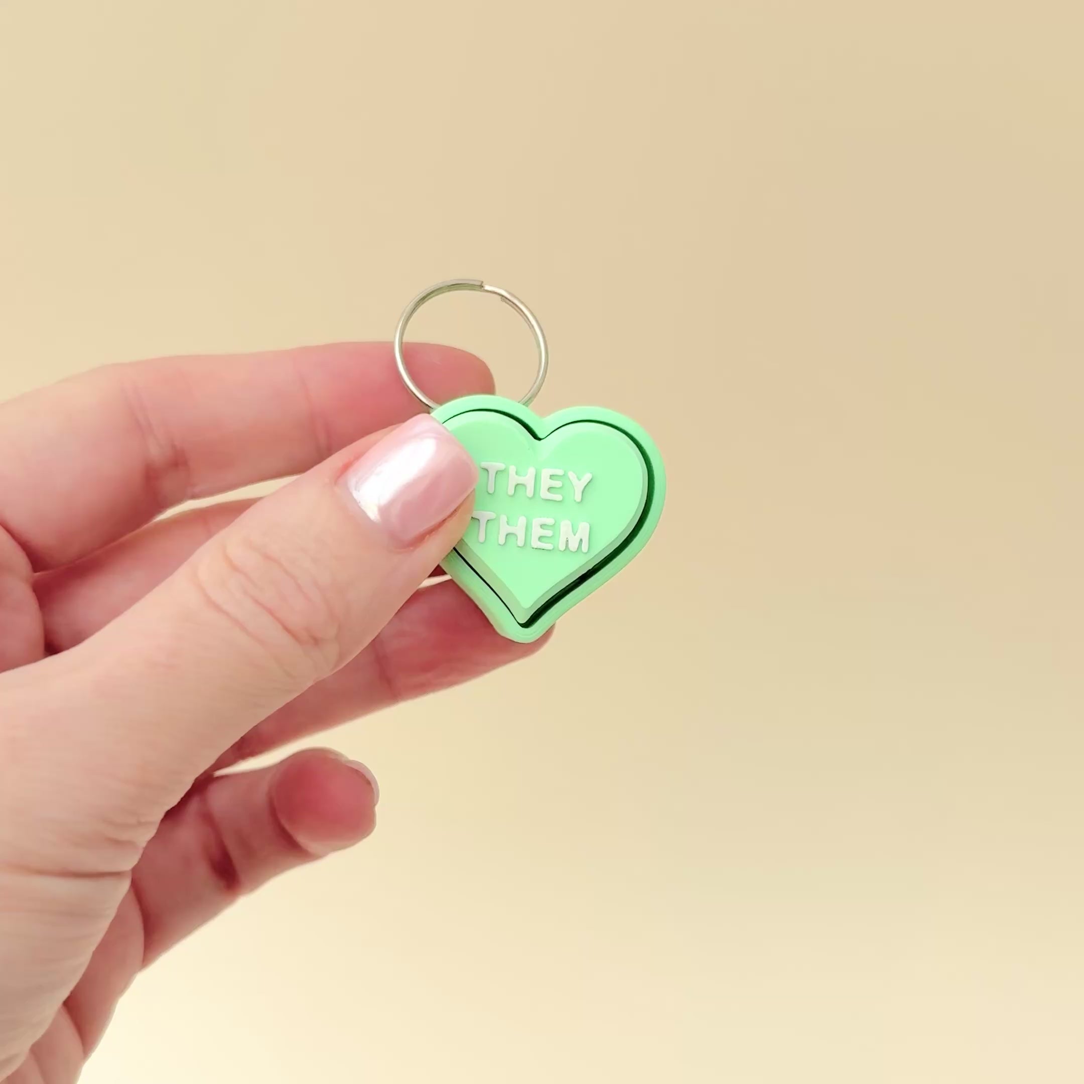 A heart-shaped mint green sensory clicker keyring available in various pronouns, including She/They.