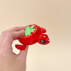 A red turtle clicker fidget toy with a green and white strawberry-style design on its shell, featuring 7 clickable spots.